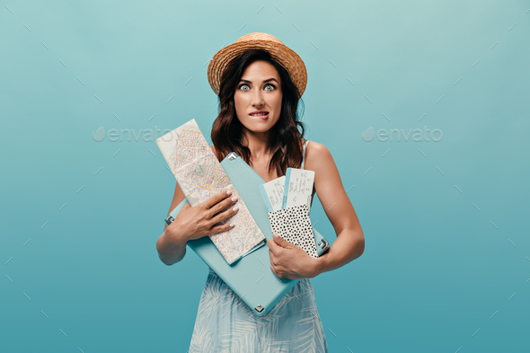 Cute girl in stylish sundress holds card, suitcase and tickets on blue background. Brunette in stra