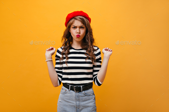 Positive woman in striped shirt and beret makes funny face. Curly young lady in denim skirt with wi