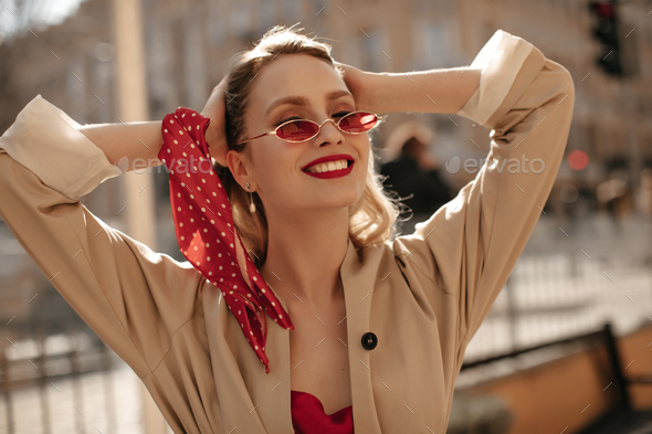 Good-humored blonde woman in beige trench coat, silk blouse and bright sunglasses smiles sincerely