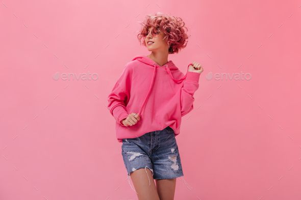 Curly woman in pink hoodie and denim shorts dance on isolated background. Happy cool girl in sungla