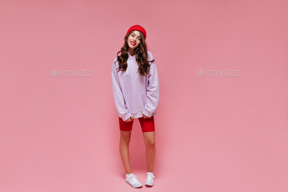Full-length portrait of brunette curly girl in red cycling shorts and purple hoodie on pink isolate