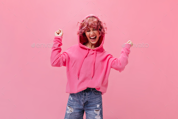Happy active woman in pink hoodie and denim pants laughing and dancing on  isolated background. Port