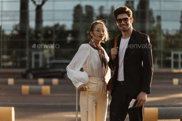 Attractive woman in eyeglasses, white blouse and beige pants poses with boyfriend outside. Portrait