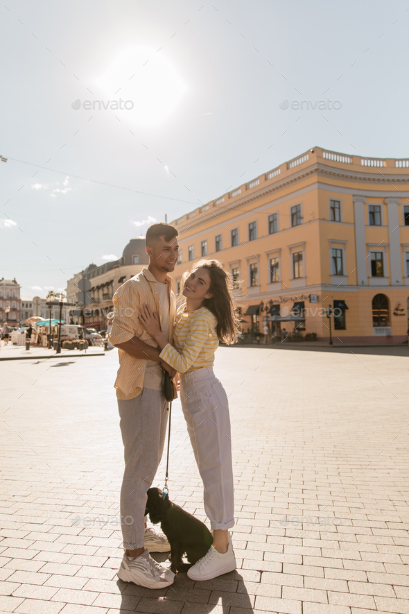 Street photo of young couple with dog in sunny city. Stylish girl wearing light pants, sneakers and