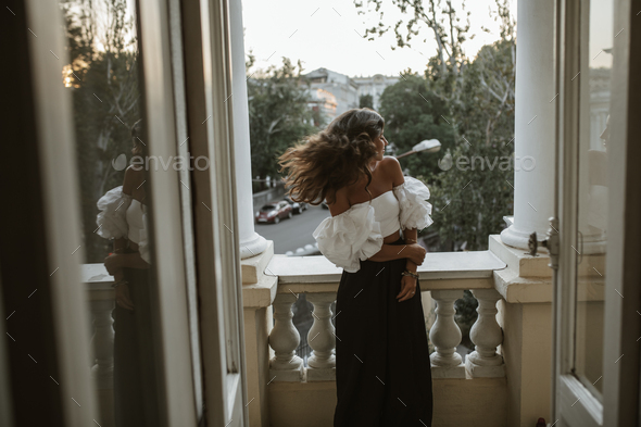 Joyful young lady with brunette wavy hair, off-shoulder top, dark pants standing on classic balcony