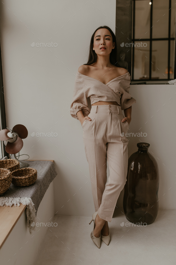 Calm elegant Asian brunette woman in beige pants and linen cropped blouse looks away and leans on w