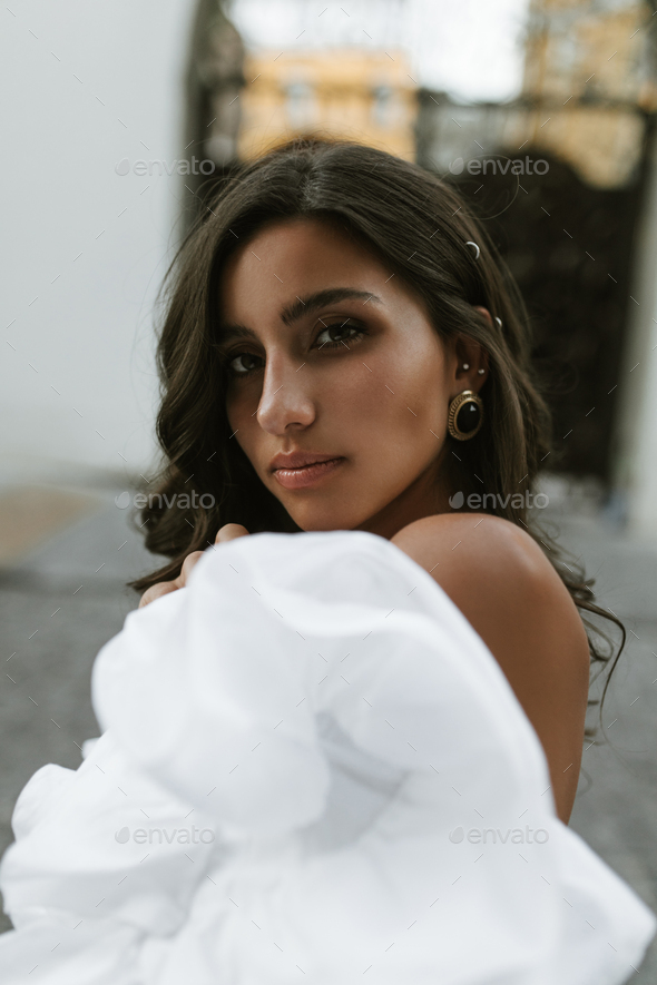 Portrait of gorgeous brunette with long wavy hair, dark eyes, tanned body in off-shoulder white top