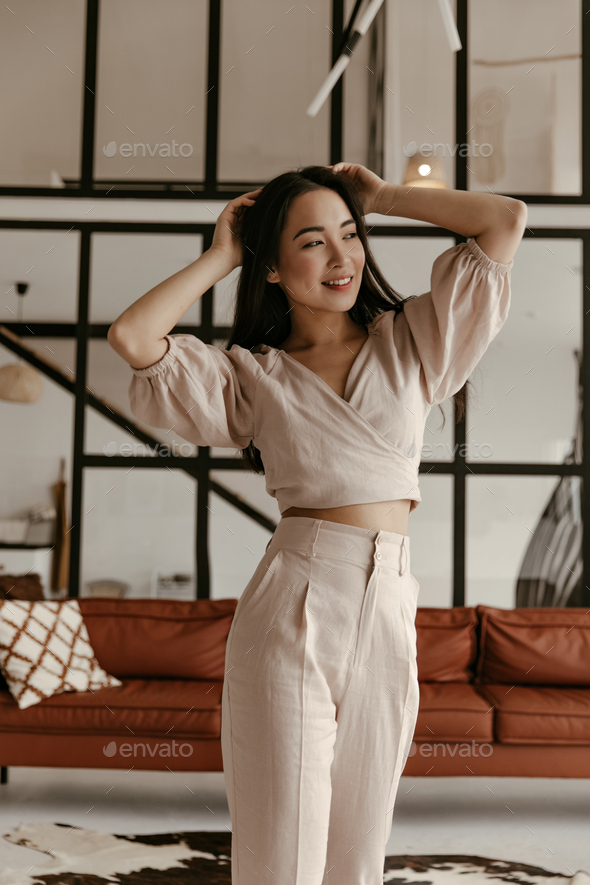 Beautiful young brunette Asian woman in stylish cropped blouse and linen beige pants touches hair,