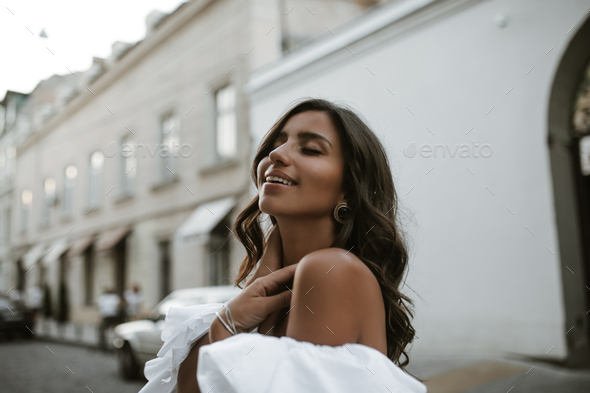 Joyful young lady with brunette wavy hair, tanned body, closed eyes in white off-shoulder blouse an