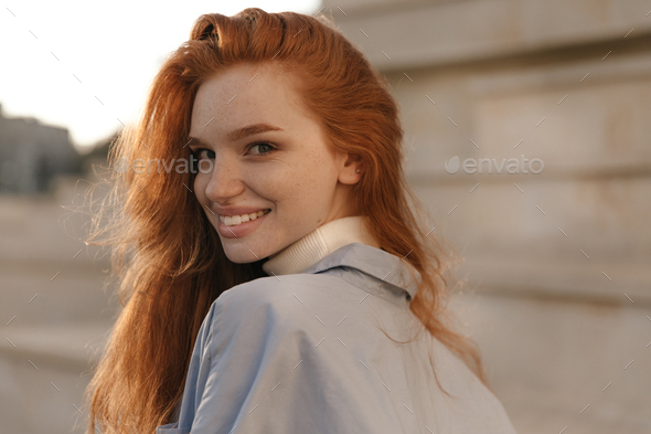 Close-up beauty portrait of young red-haired girl smiling and turning back outdoors. Pretty ginger