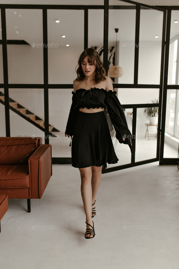 Pretty brunette woman in black cropped top and skirt walks in cozy stylish living room. Charming la