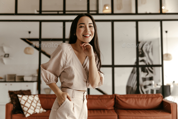 Pretty brunette Asian woman in cropped beige top and linen pants laughs, looks into camera and pose