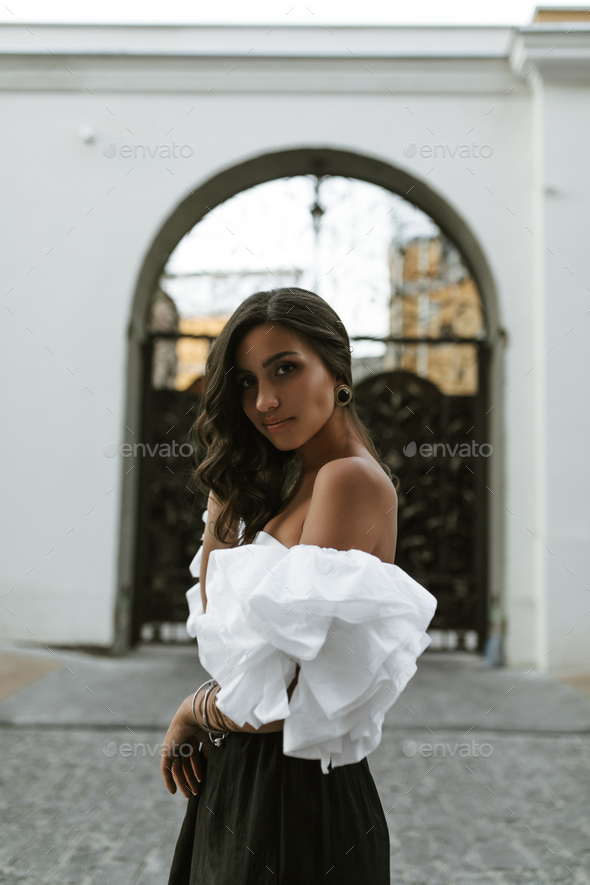 Charming young brunette with curly hair, dark eyes and slim tanned body in white off-shoulder top,