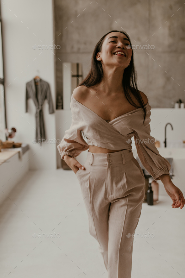 Joyful brunette tanned Asian woman in cropped beige blouse and linen pants moves in cozy bathroom a