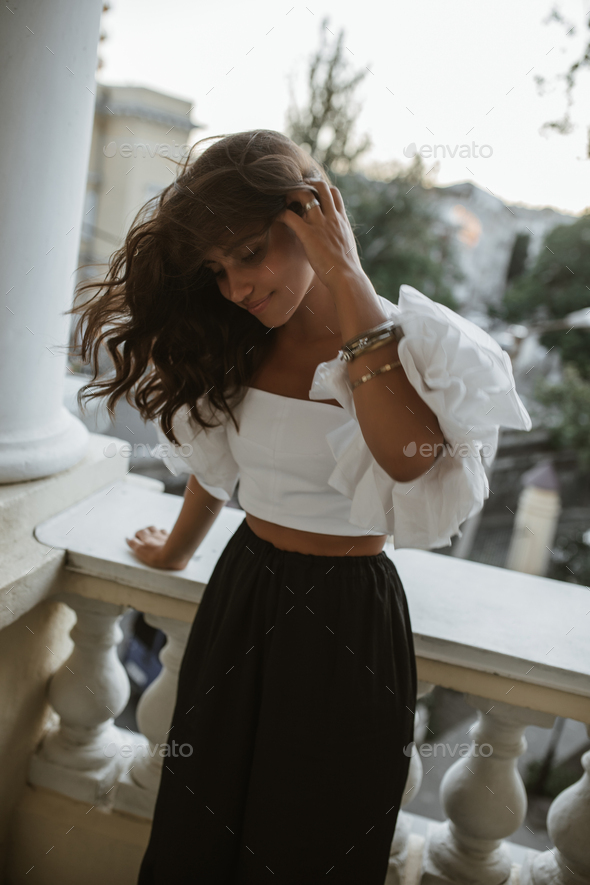 Cute young brunette in white off-shoulder top, dark high-wasted pants and stylish accessories stand