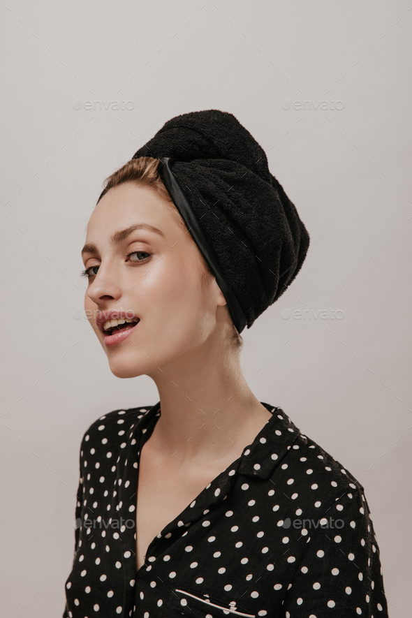 Excited young woman in black polka dot shirt and silk towel on head, looking into camera against li