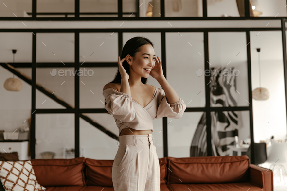 Joyful brunette Asian woman in cropped top and beige linen pants smiles, touches hair and poses in