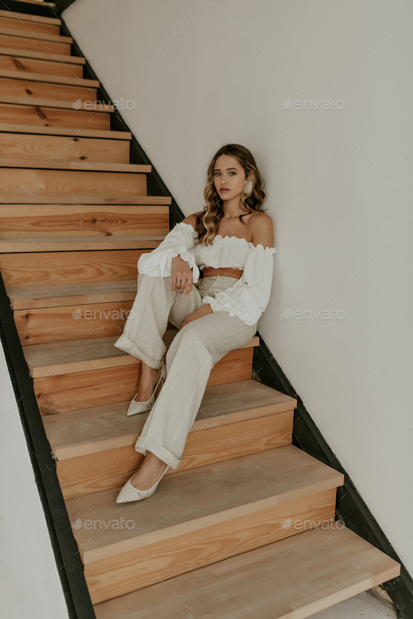 Curly lady in cropped white blouse, beige linen pants and high heels looks into camera, leans on wa