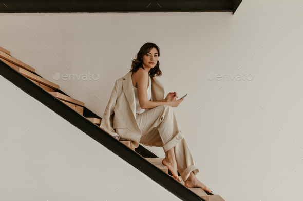 Young brunette curly woman in beige suit sits on wooden stairs. Pretty tanned lady in linen pants a