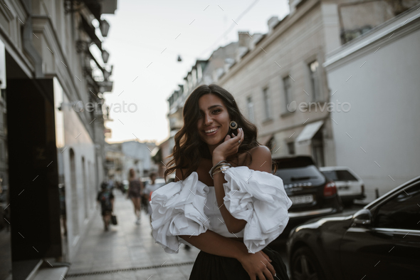 Joyful lady with brunette curly hair and tanned body, in retro outfit of white off-shoulder blouse,