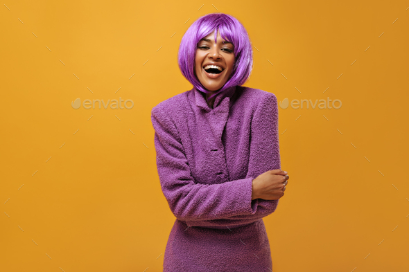 Happy woman with wig in warm purple outfit smiling on isolated backdrop. Short-haired lady in lilac