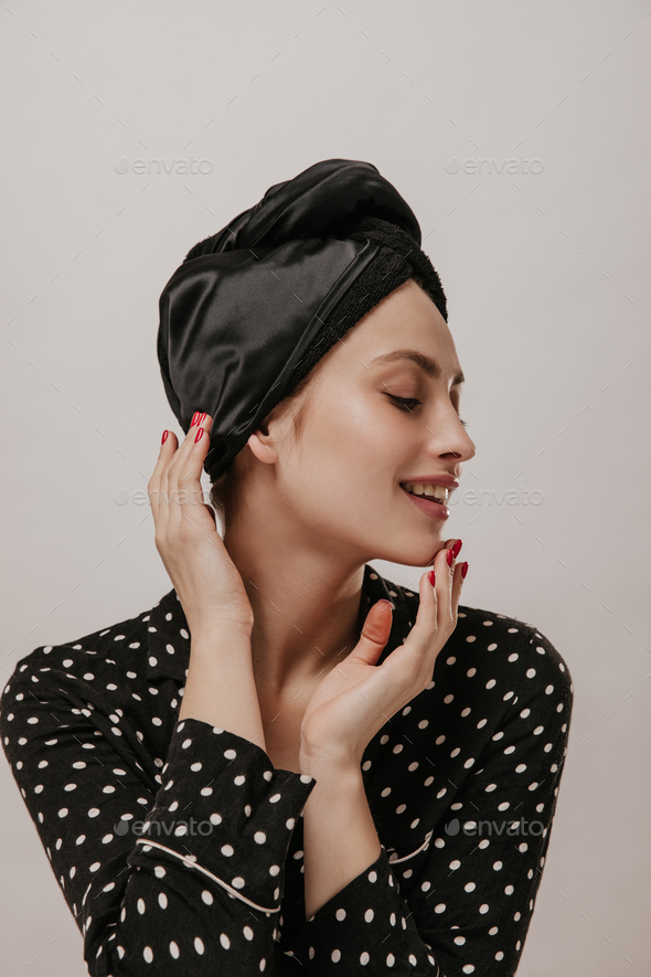 Gorgeous young lady in black silk towel on head and polka dot pajama shirt touching face, smiling a