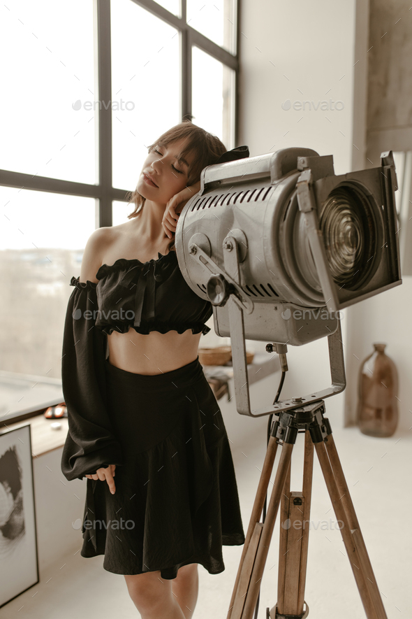 Pretty brunette woman in stylish skirt and black cropped blouse leans on huge floor lamp and poses