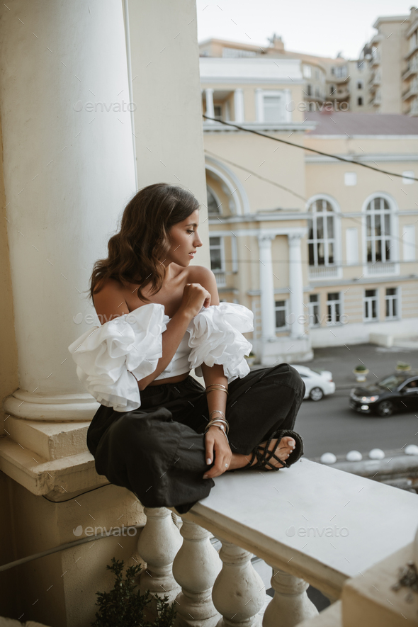 Engaging young brunette in white off-shoulder blouse, black pants and sandals, sitting on classic s