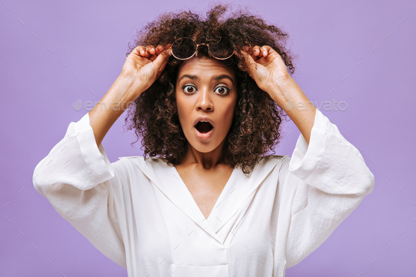 Shocked girl in light blouse takes off glasses on lilac background. Brown-eyed lady with curly hair