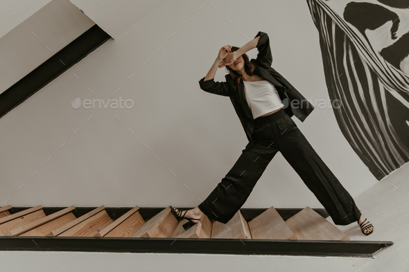 Photo of brunette curly woman in loose black pants and oversized jacket stands in strange pose on w