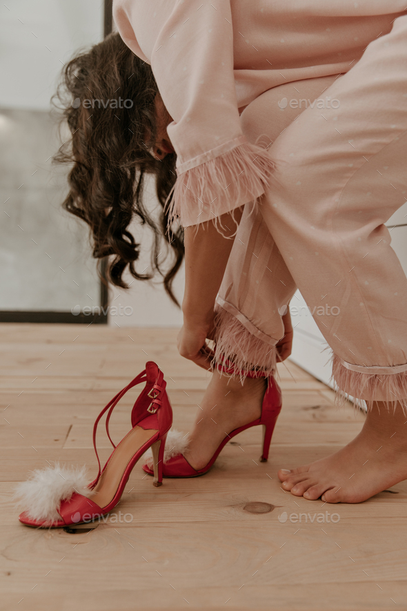 Curly brunette woman in pink polka dot home suit puts in red high heels with feathers. Portrait of