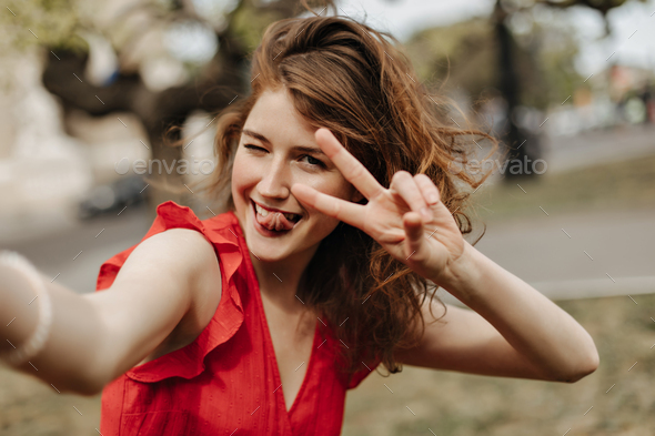 Cheerful woman with wavy hair in bright clothes showing tongue and peace sign outside. Girl in red