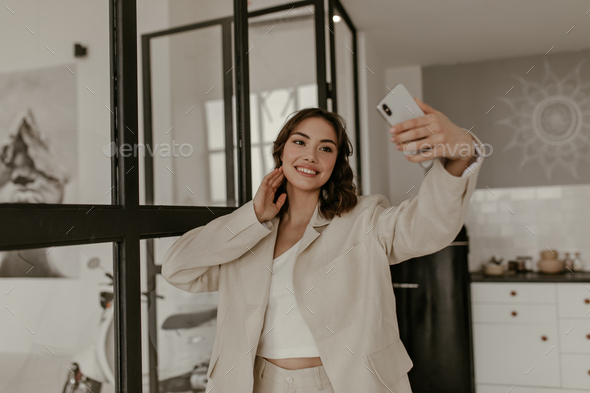 Brown-eyed brunette curly woman in beige loose jacket and linen pants smiles sincerely, holds phone