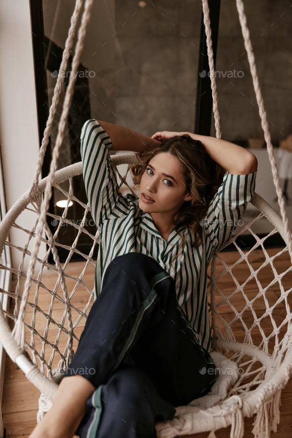 Relaxed attractive woman in striped silk shirt and pajama pants sits in comfortable wicked chair an