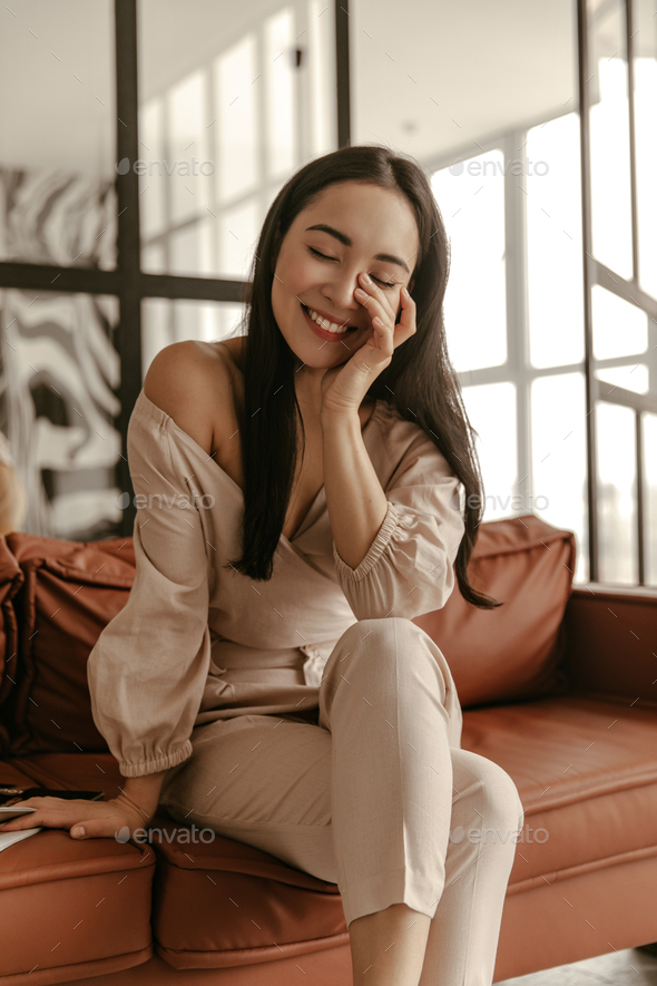 Joyful brunette Asian woman in stylish beige cropped top and linen pants smiles sincerely with clos