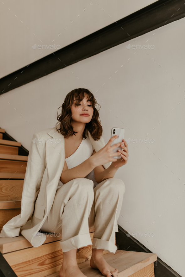 Curly charming brunette woman in stylish linen pants, beige jacket and white top holds phone and si