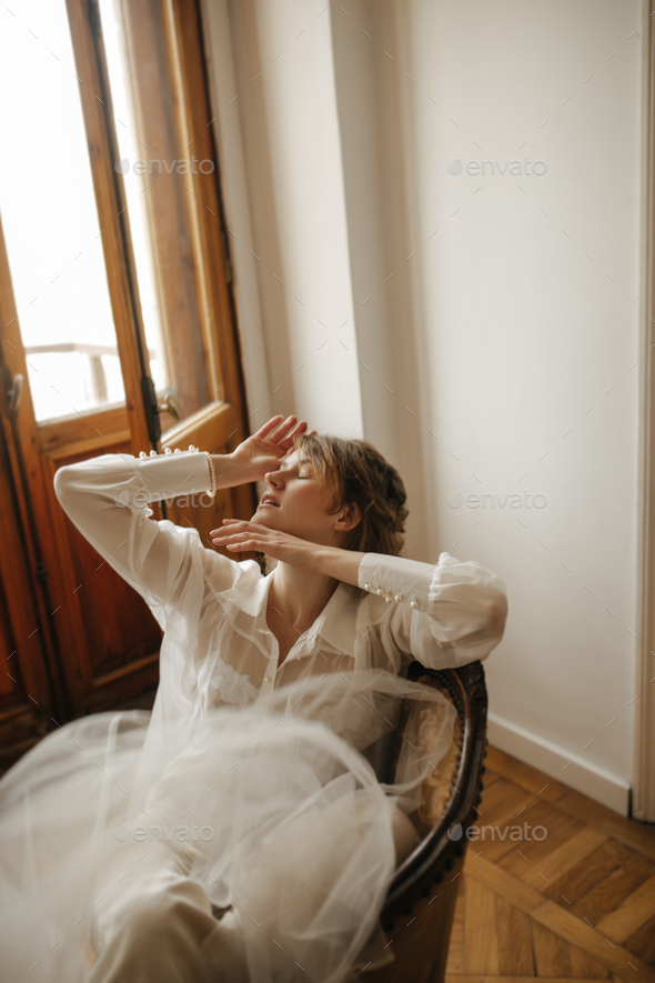Modern lady in light blouse sitting in armchair inside. Cool woman transparent blouse posing with p