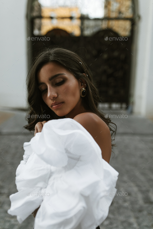 Tender brunette with long curly hair, smooth tanned body in white off-shoulder top, vintage earring