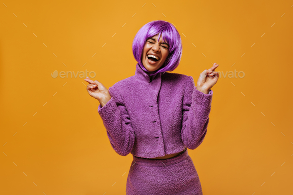 Optimistic girl in bright wig showing peace sign on yellow backdrop. Modern woman in lilac jacket l