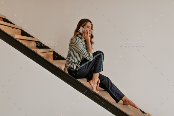 Beautiful curly long-haired woman in silk black pajama pants and striped shirt talks on cellphone a