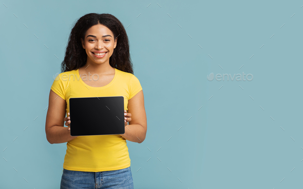 Cool website. Happy black lady showing tablet with blank screen, standing over blue studio