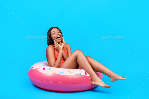 Portrait of excited black lady in bikini touching her face, sitting in donut shaped inflatable ring
