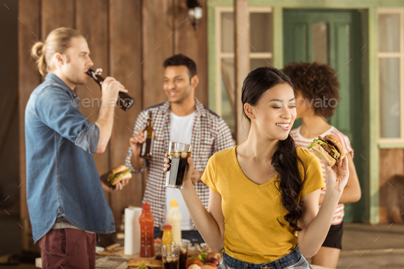 young asian girl eating burger and drinking cola with friends at picnic on patio