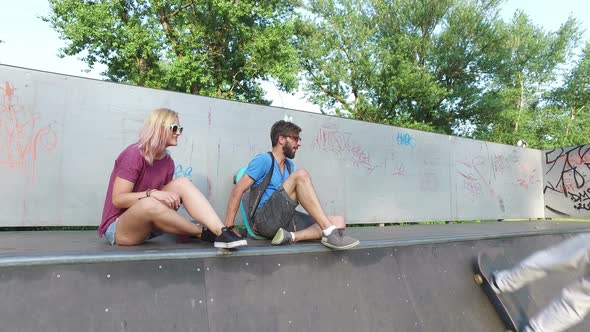 Man and woman sitting on ramp at skatepark, watching their friend skateboarding alt