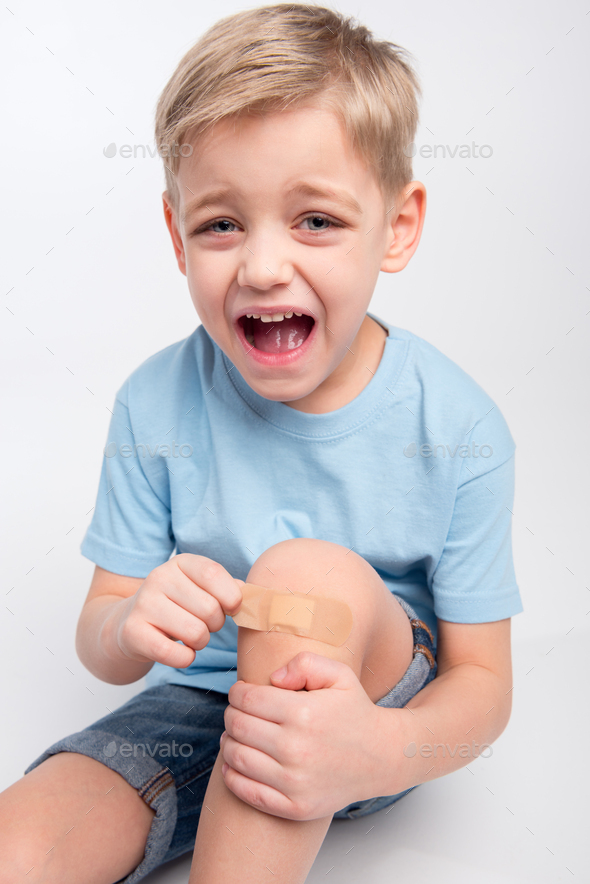 Little boy removing patch from injured knee and crying at camera