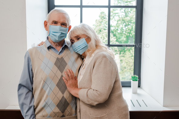 upset elderly couple in medical masks hugging at home during self isolation