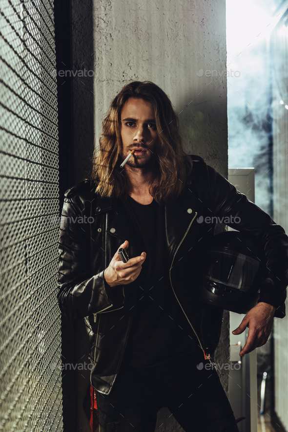 Handsome long haired man in leather jacket holding helmet and lighting cigarette with lighter