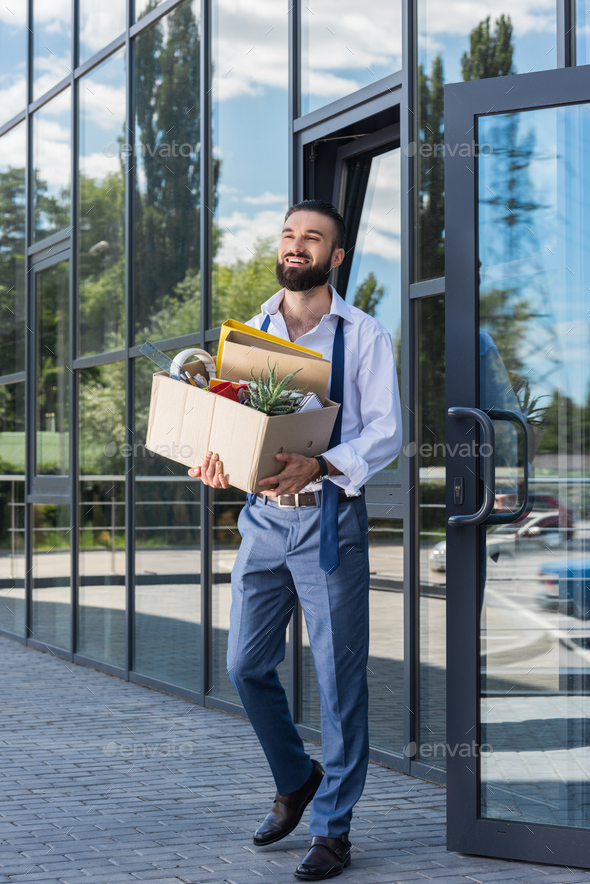 happy businessman with cardboard box walking out office building, quitting job concept