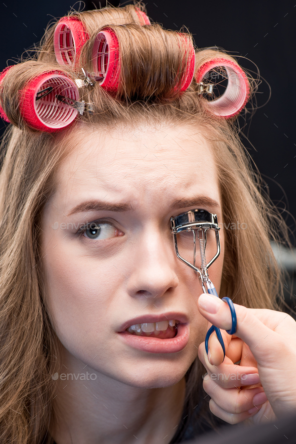 Makeup artist correcting eyelashes with curling tongs of scared young woman in curlers