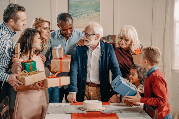 large family presenting cake and gifts to grey haired grandfather